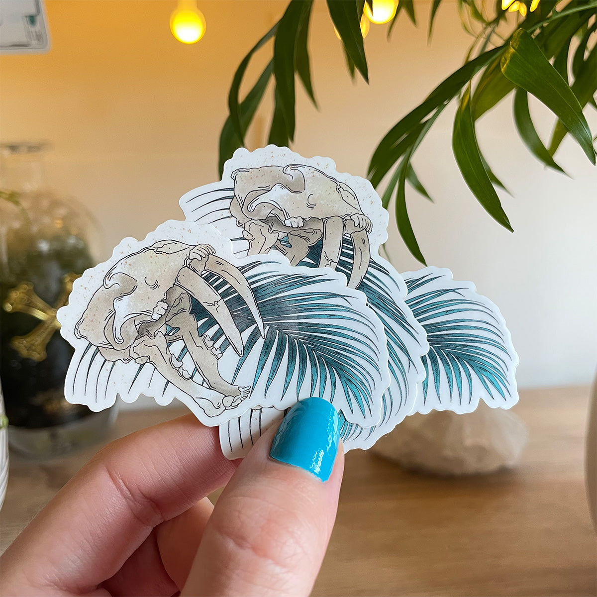 hand holding several stickers of sabertooth skull with open mouth single palm leaf behind.