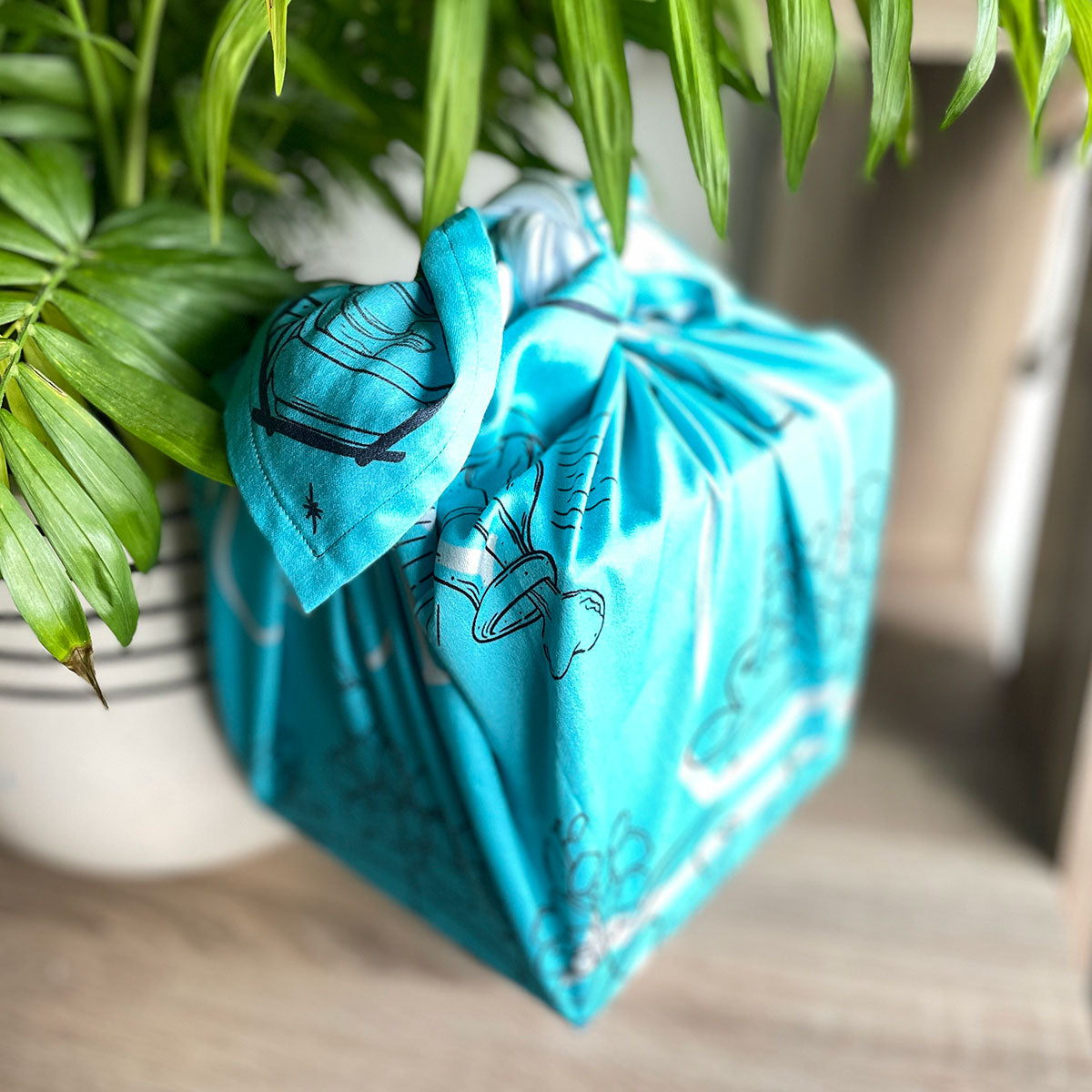Square fabric wrapped and tied around cube gift box, turquoise bandana in satin cotton illustrated with unique design, floating next to plant vase