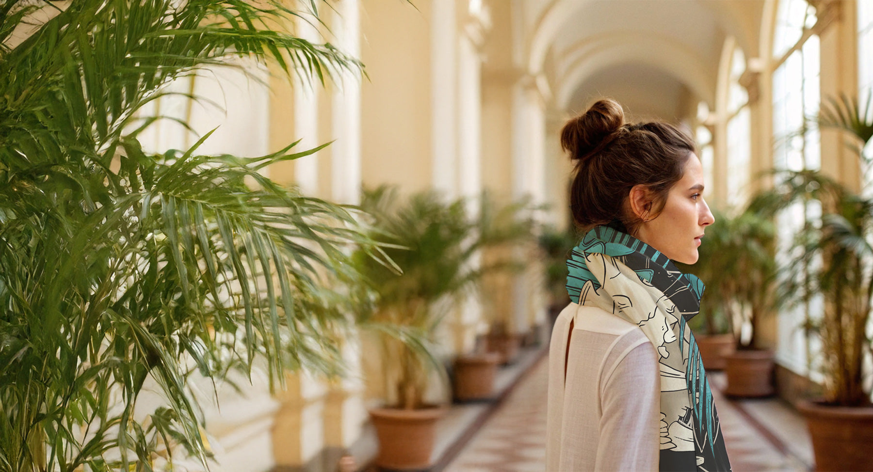 Woman walking through a sunlit hallway with plants on both sides; she wears a printed scarf of an animal skull in black and teal wrapped around the neck
