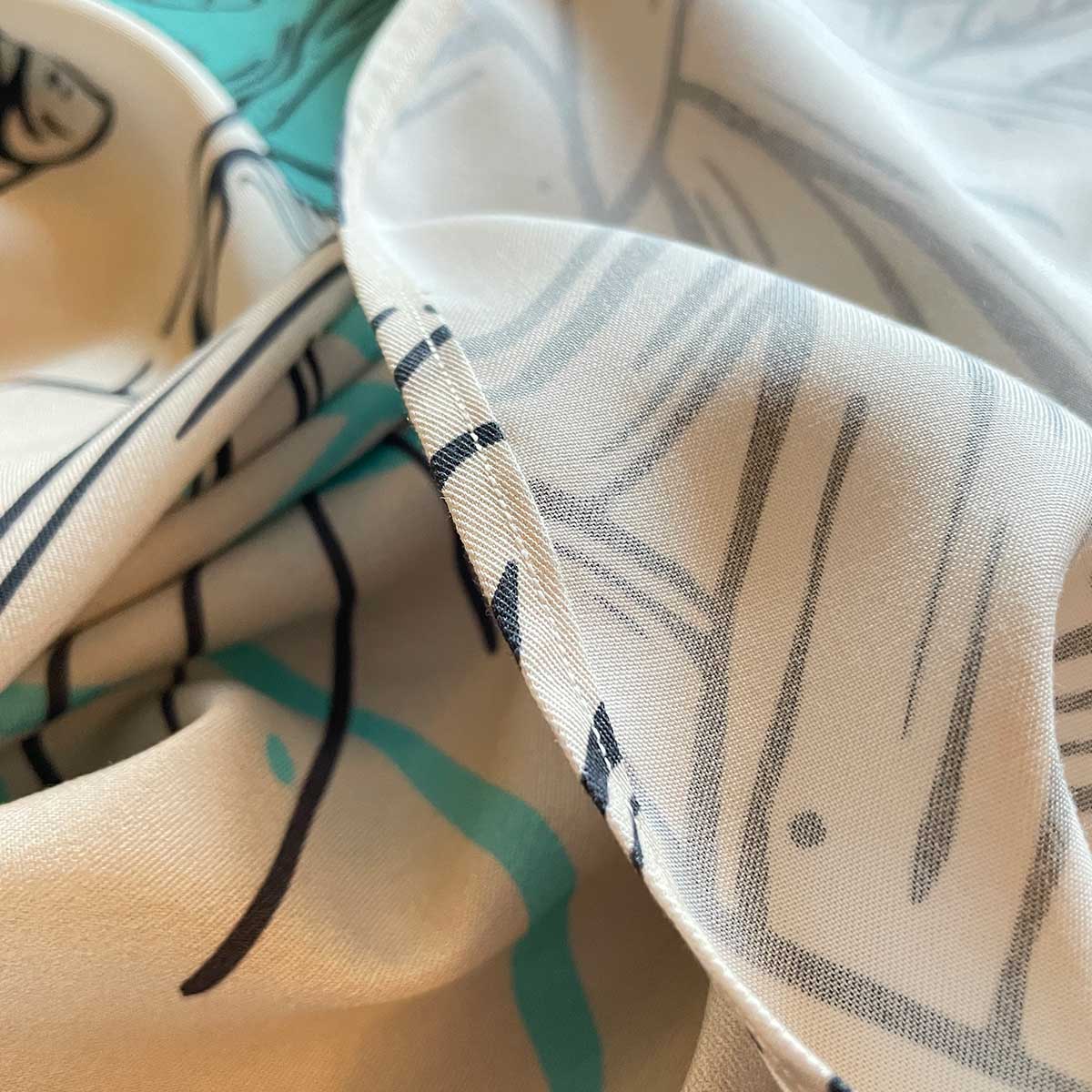 Close up of folded, sewed hem of fabric, front and back of fabric in cream, turquoise and black outlined drawings of plants and leaves