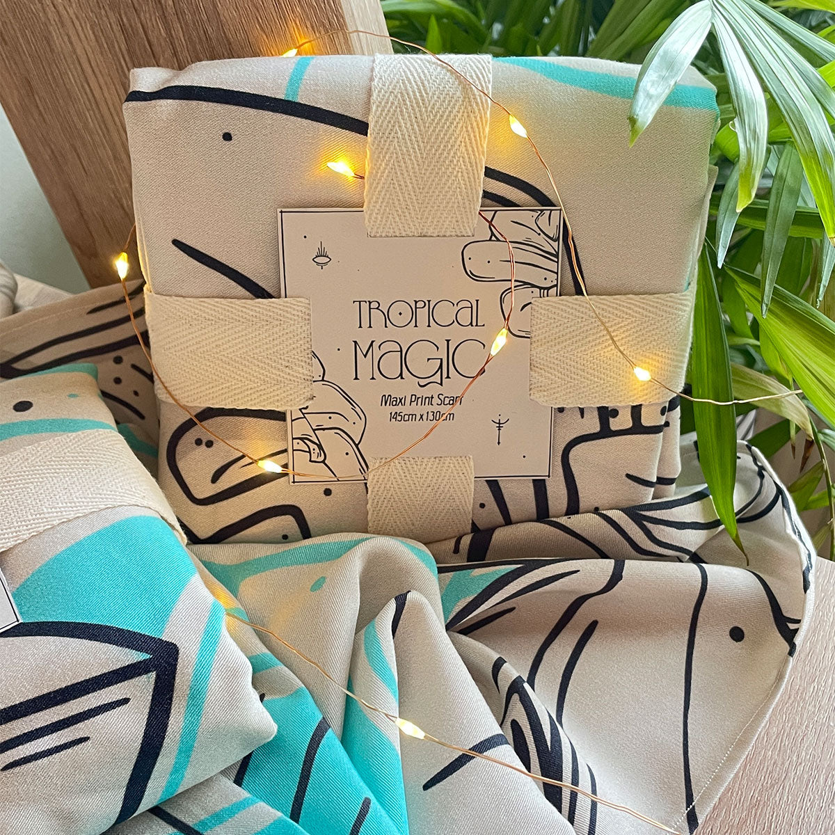 Fabric folded in square tied with cotton ribbon and square paper with description. Sitting on table, palm leaves next, sunlit room, fairy lights surround it. Another fabric sits under it.