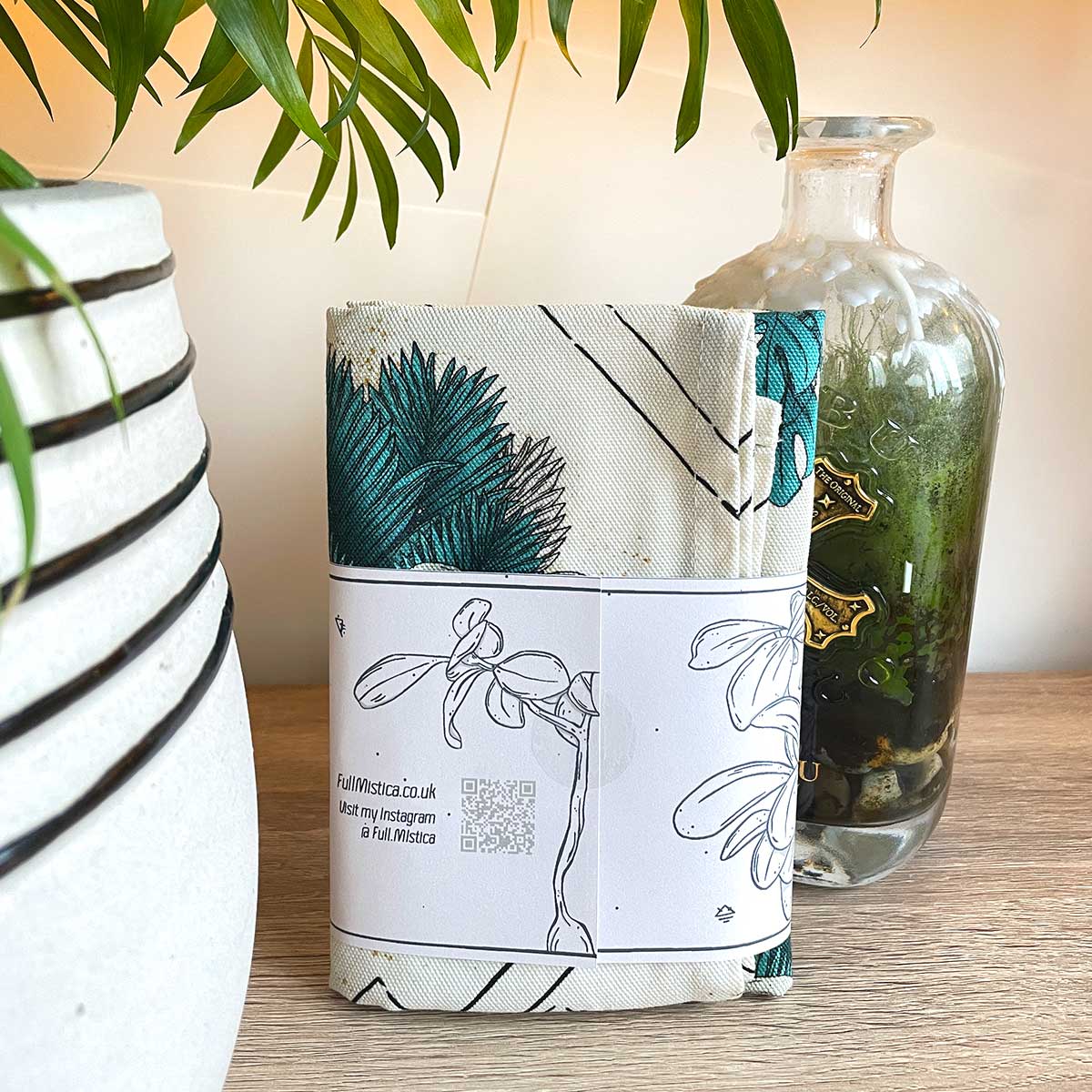 Tea towel with belly band standing on wooden table. Plant pot on the left. Tropical leaves in teal with bird skull design, runes and lines in black.