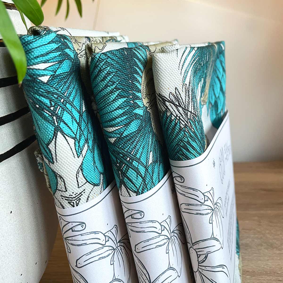 Three tea towels sideways with belly band standing on plant pot to the left. Designs of tropical leaves in teal with lion skull open mouth