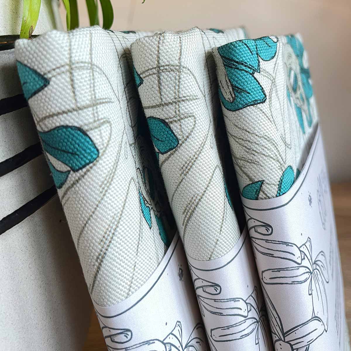 Three tea towels sideways with belly band standing on plant pot on the left. Designs of monstera leaves in teal with bird skull outline leaves as watermark in background