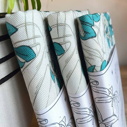 Three tea towels sideways with belly band standing on plant pot on the left. Designs of monstera leaves in teal with bird skull outline leaves as watermark in background