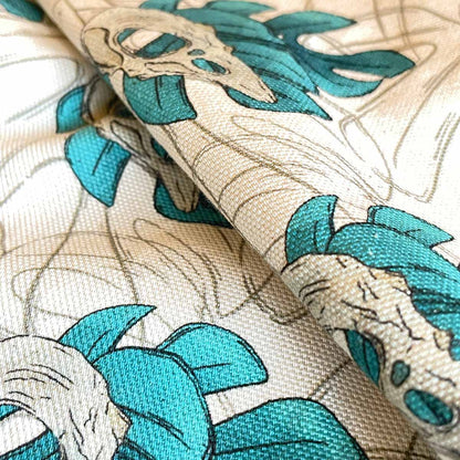 Close up of tea towel folded fabric. Monstera leaves in teal with bird skull design, outline leaves watermark in background, all over pattern