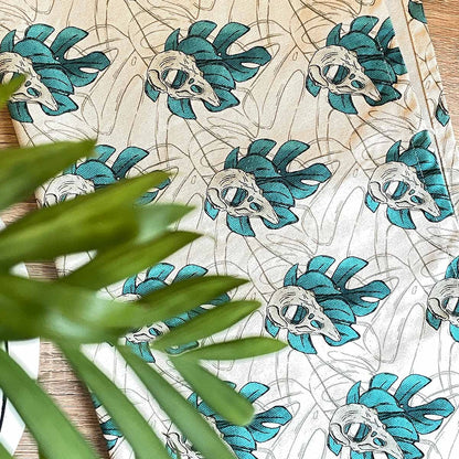 Tea towel open top view. Monstera leaves in teal with bird skull design, outline leaves watermark in background, all over pattern