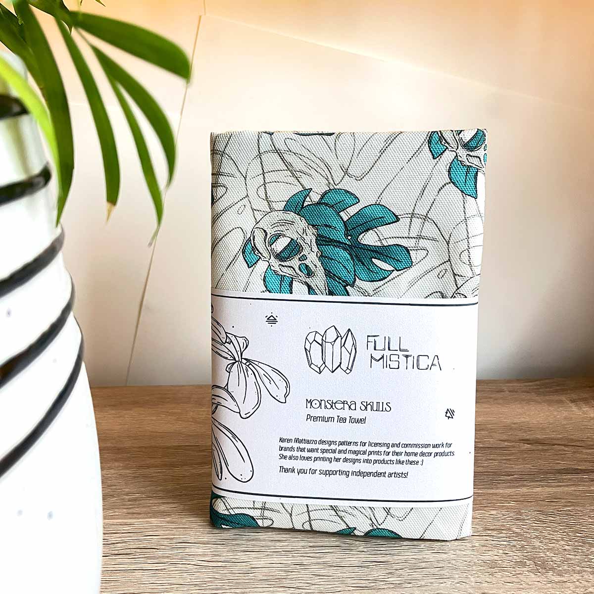 Tea towels front with belly band standing on wooden table plant pot on the left. Designs of monstera leaves in teal with bird skull outline leaves as watermark in background