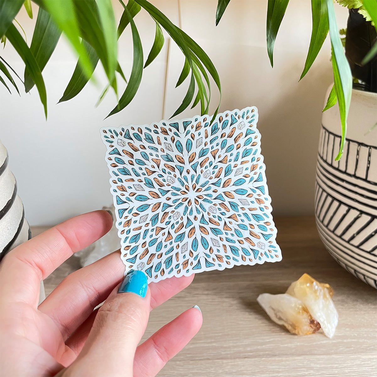 hand holding sticker of several tiny leaves and small gemstones in teal and orange organised like a tile shaped like a square