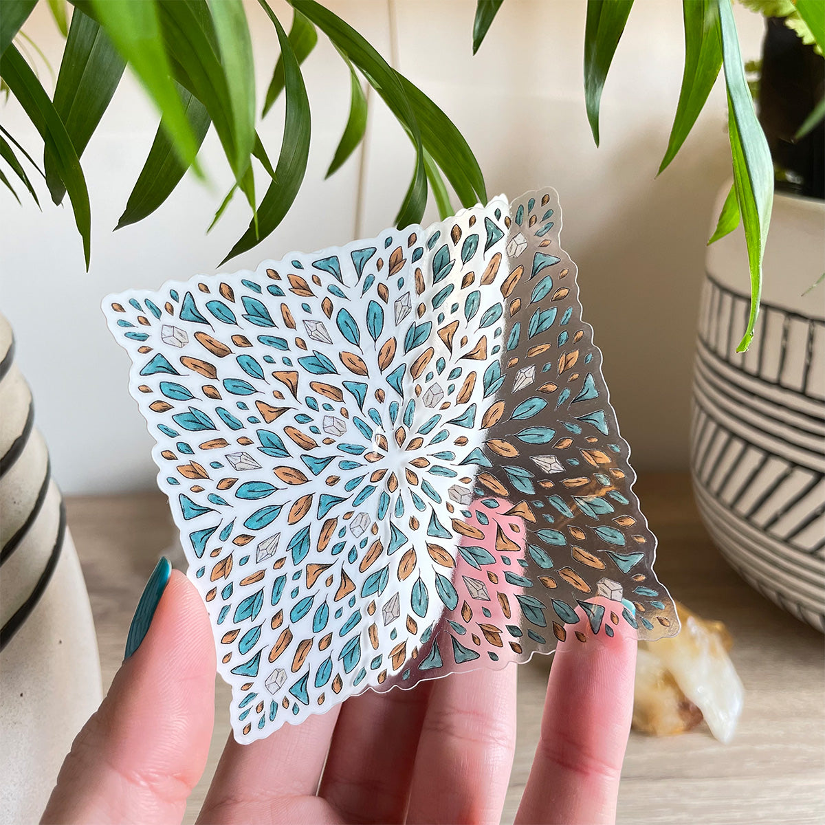 hand holding clear sticker of several tiny leaves and small gemstones in teal and orange organised like a tile shaped like a square