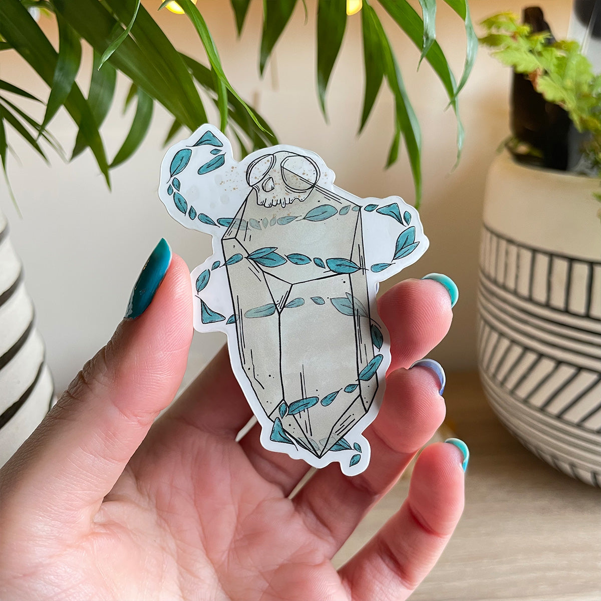 hand holding sticker of big quartz crystal with tiny monkey skull on top, swirls of teal leaves floating around it