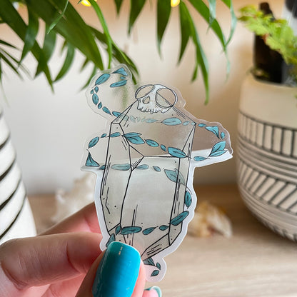 hand holding clear transparent sticker of big quartz crystal with tiny monkey skull on top, swirls of teal leaves floating around it