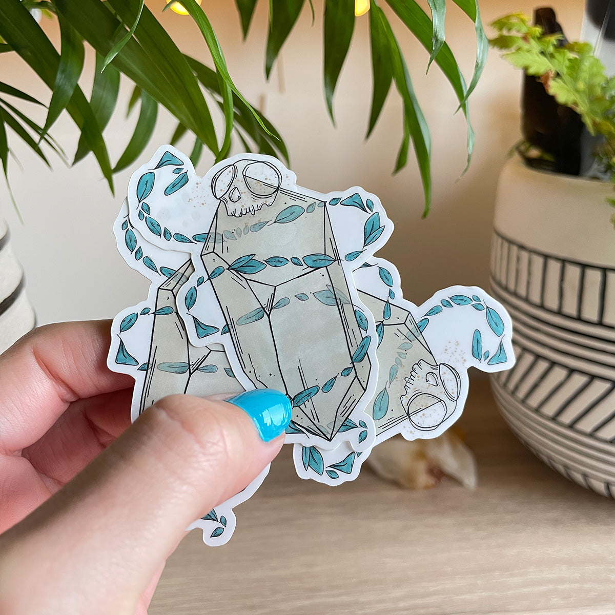 hand holding several stickers of big quartz crystal with tiny monkey skull on top, swirls of teal leaves floating around it