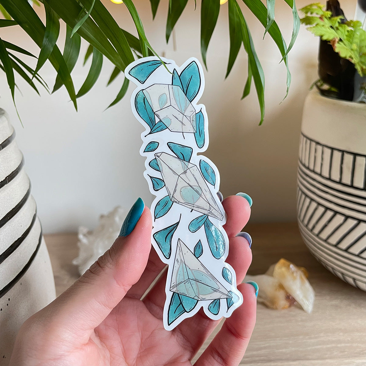 Hand holding clear sticker trio of different clear quartz crystals with loads of teal leaves behind. Background with two plant pots