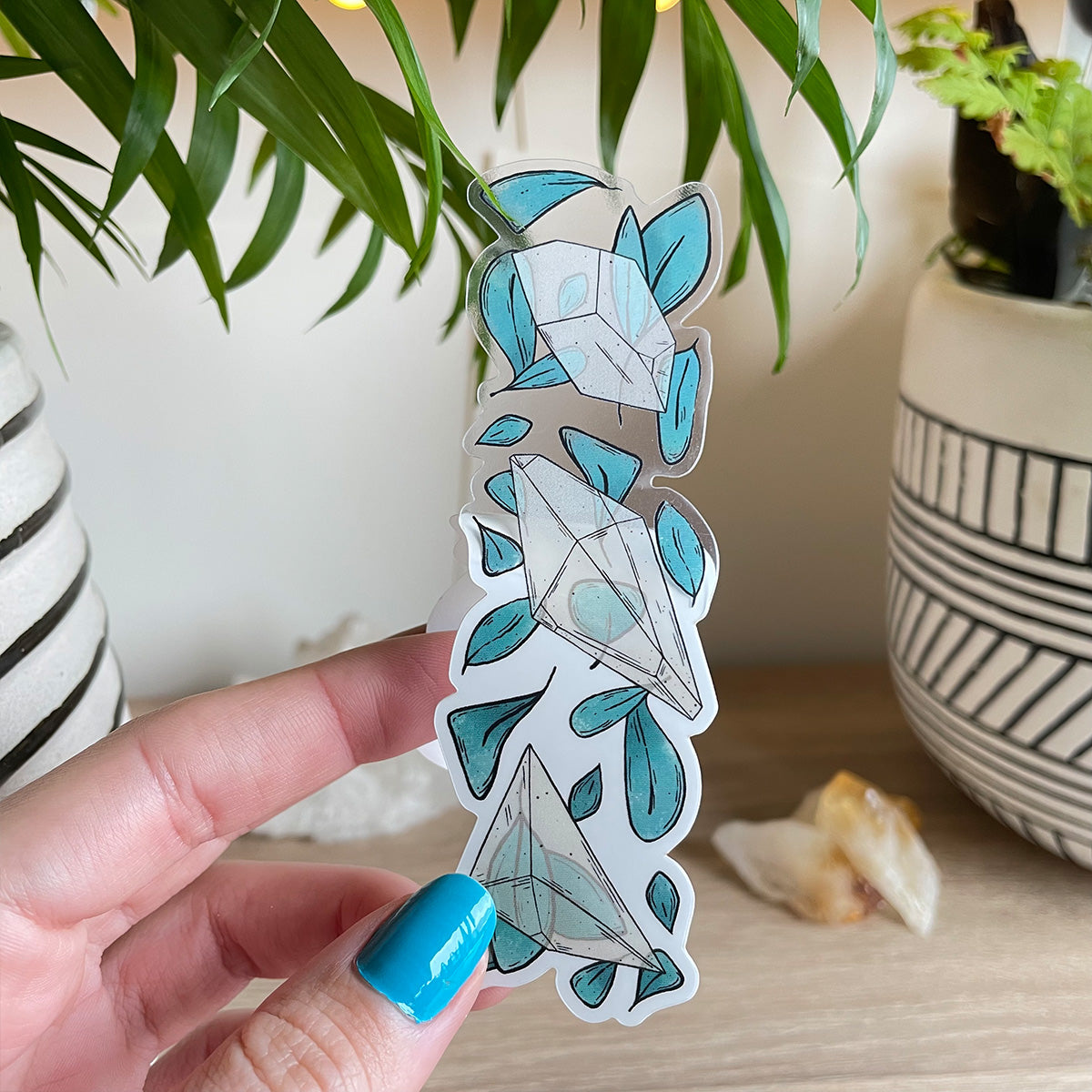 Hand holding clear sticker trio of different clear quartz crystals with loads of teal leaves behind. Background with two plant pots