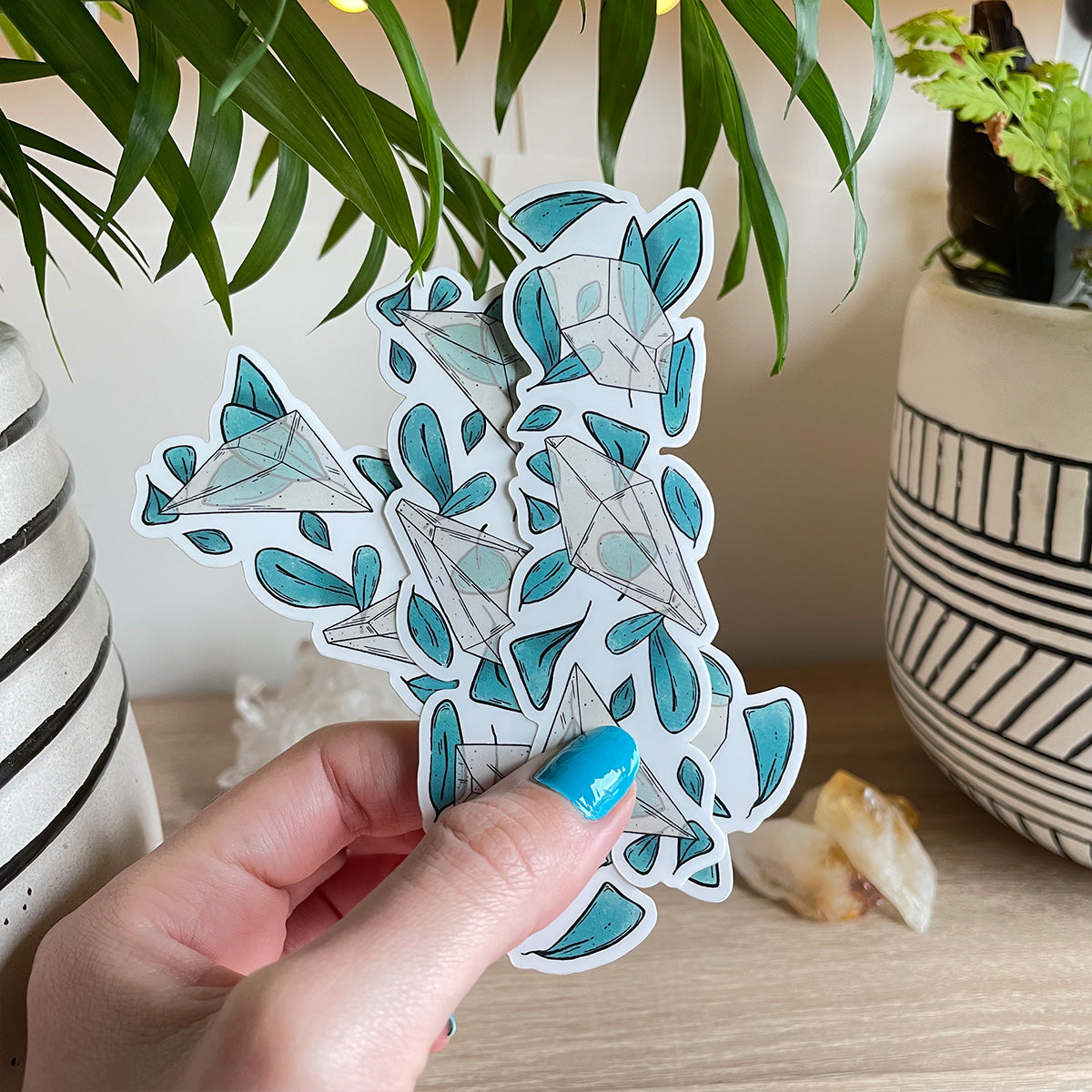 Hand holding several clear stickers trio of different clear quartz crystals with loads of teal leaves behind. Background with two plant pots