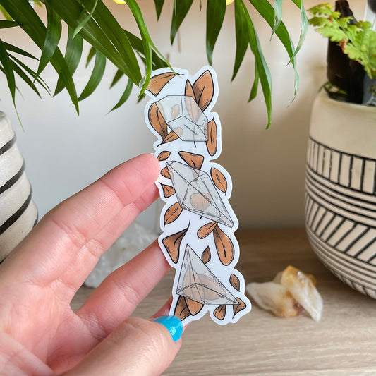 Hand holding clear sticker trio of different clear quartz crystals with loads of orange leaves behind. Background with two plant pots