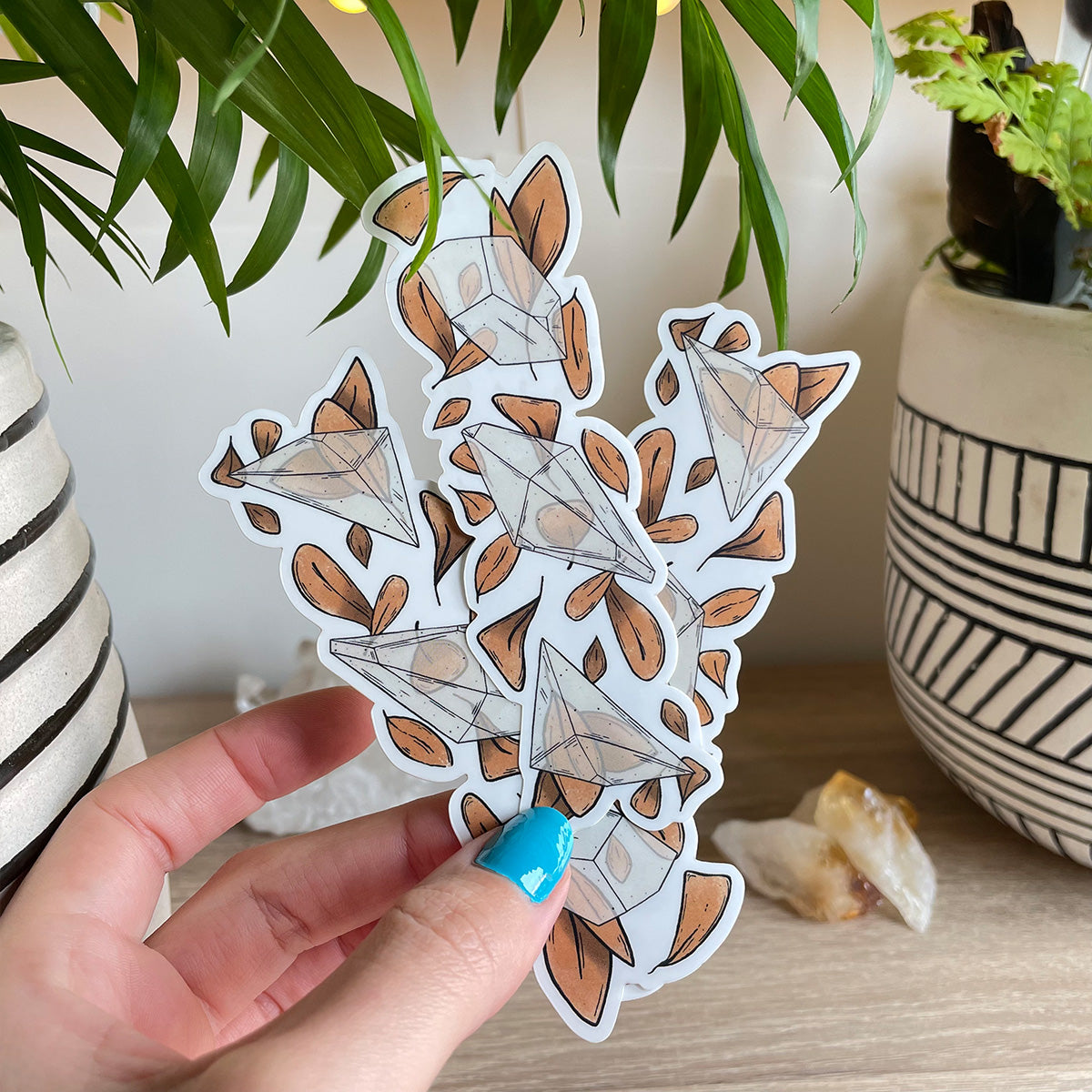 Hand holding several clear sticker trio of different clear quartz crystals with loads of orange leaves behind. Background with two plant pots