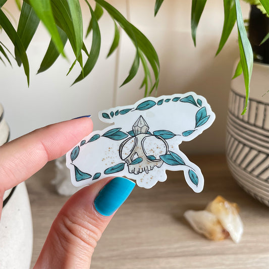 Hand holding sticker of tiny skull with crystal and several leaves floating around. Background has two plant pots and citrines sitting on table