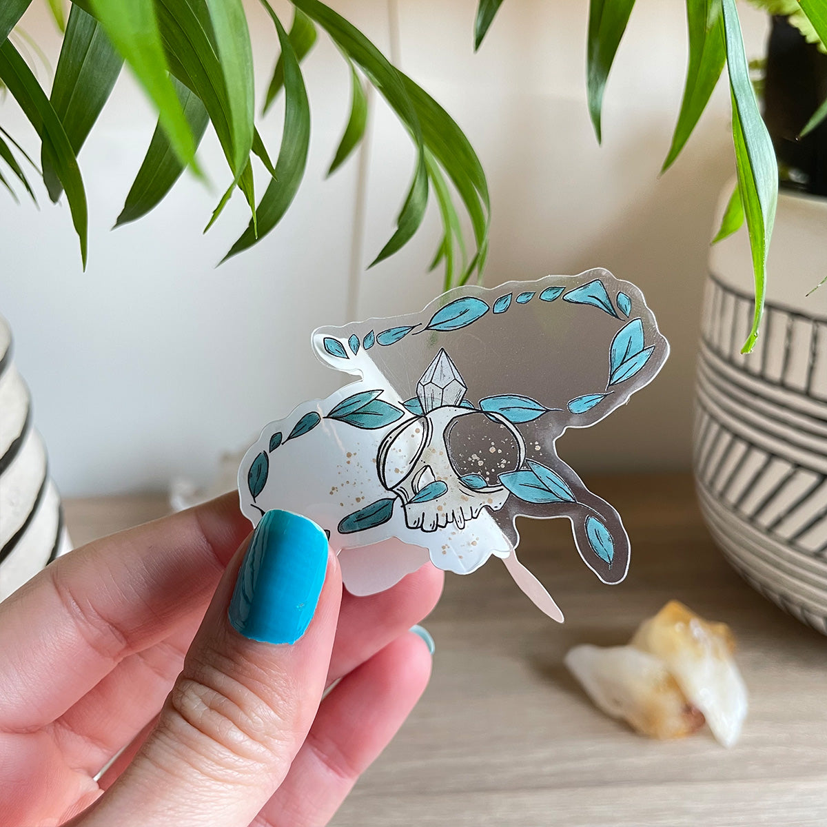 Hand holding clear sticker of tiny skull with crystal and several leaves floating around. Background has two plant pots and citrines sitting on table