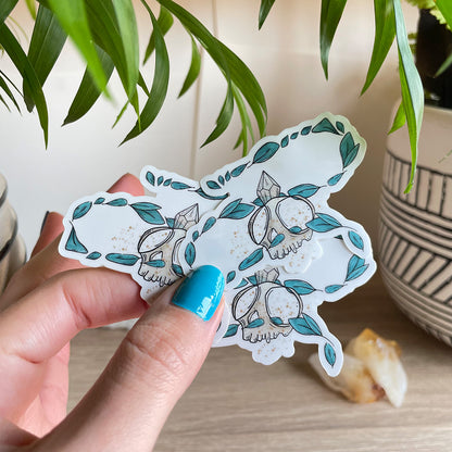 Hand holding several stickers of tiny skull with crystal and several leaves floating around. Background has two plant pots and citrines sitting on table