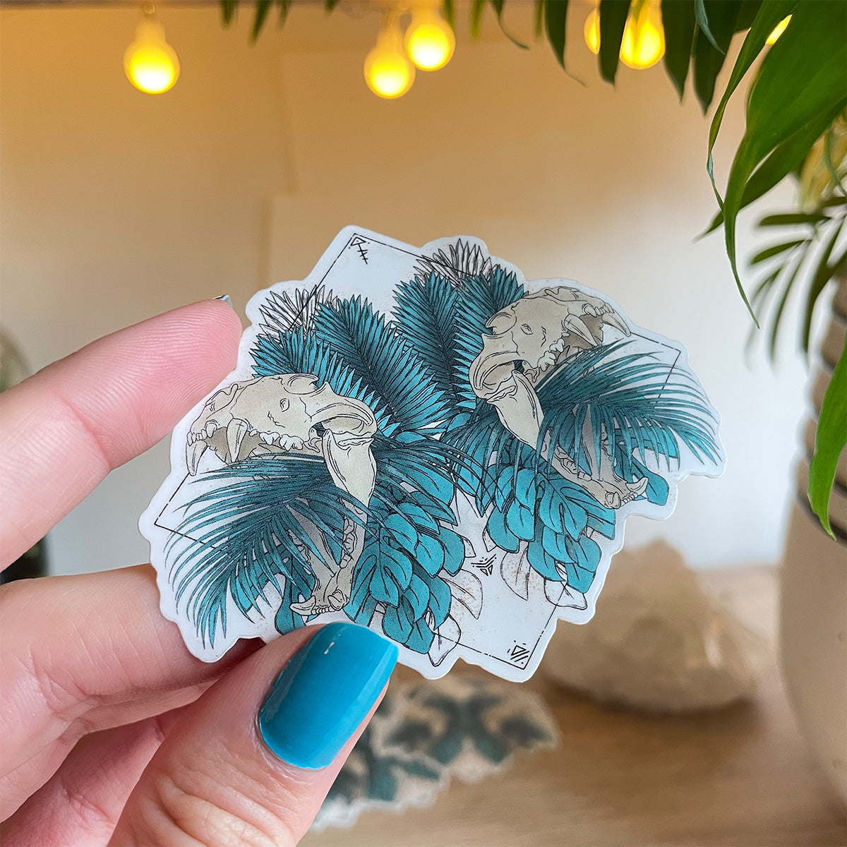 Hand holding sticker, designs of tropical leaves in teal with two lion skulls open mouths mirrored, runes and black lines in wide diamond shape