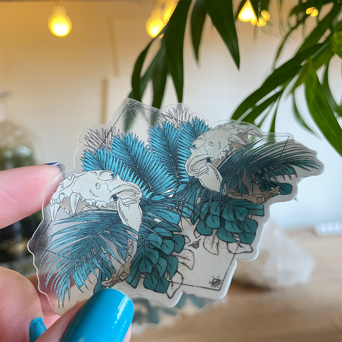 Hand holding clear transparent sticker, design of tropical leaves in teal with two lion skulls open mouths mirrored, runes and black lines in wide diamond shape