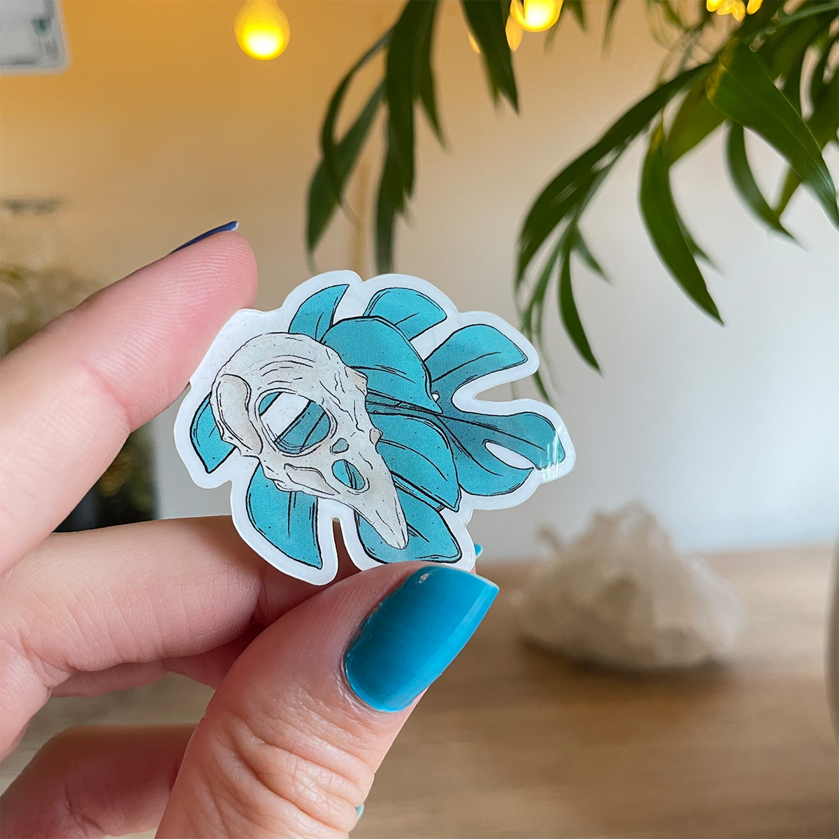 hand holding tiny sticker of two monstera leaves in teal, bird skull above leaves, big crystal and palm leaves blurry behind