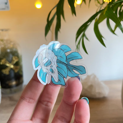 hand holding tiny clear transparent sticker of two monstera leaves in teal, bird skull above leaves, big crystal and palm leaves blurry behind