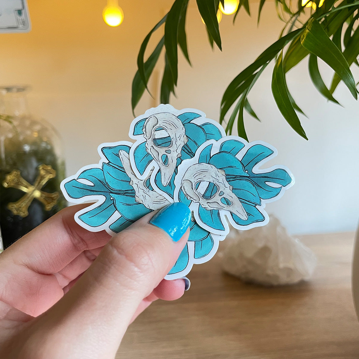 hand holding several tiny stickers of two monstera leaves in teal, bird skull above leaves, big crystal and palm leaves blurry behind