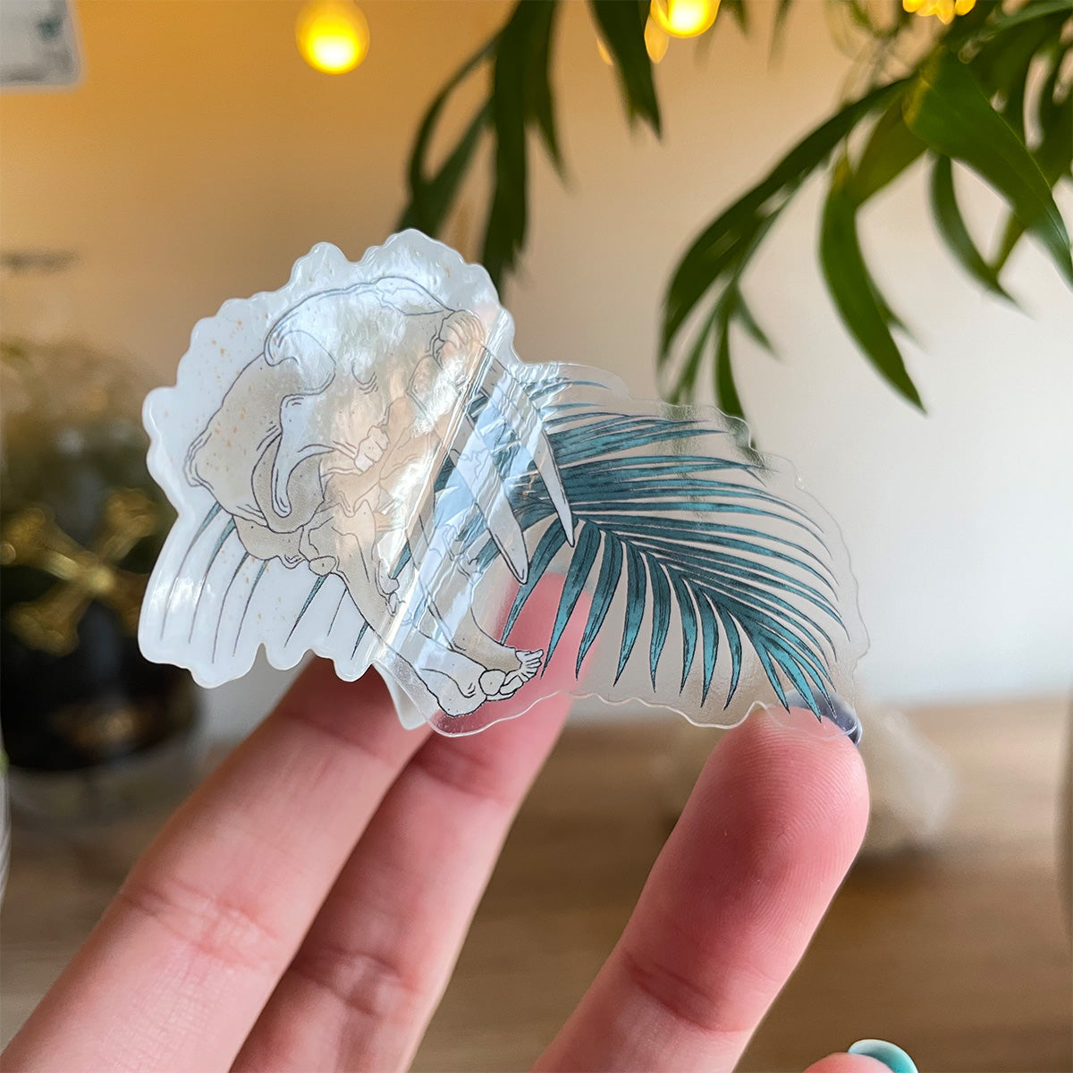 hand holding clear sticker of sabertooth skull with open mouth single palm leaf behind. Background with palm leaf and cozy dotted lights blurry