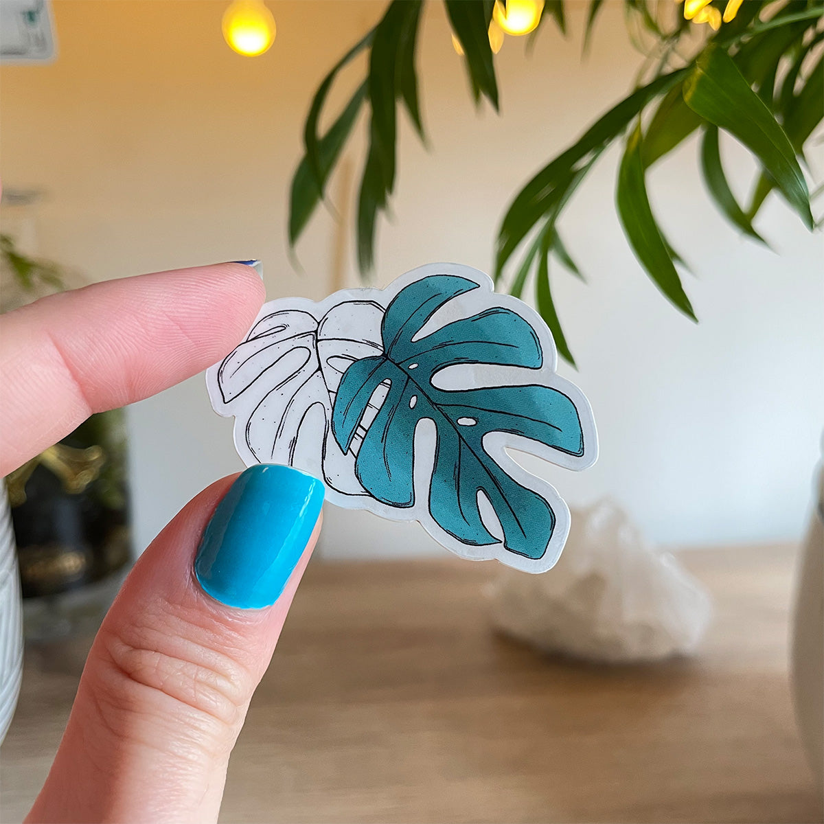 Hand holding sticker of two monstera leaves, one is outlined in black, the other teal coloured