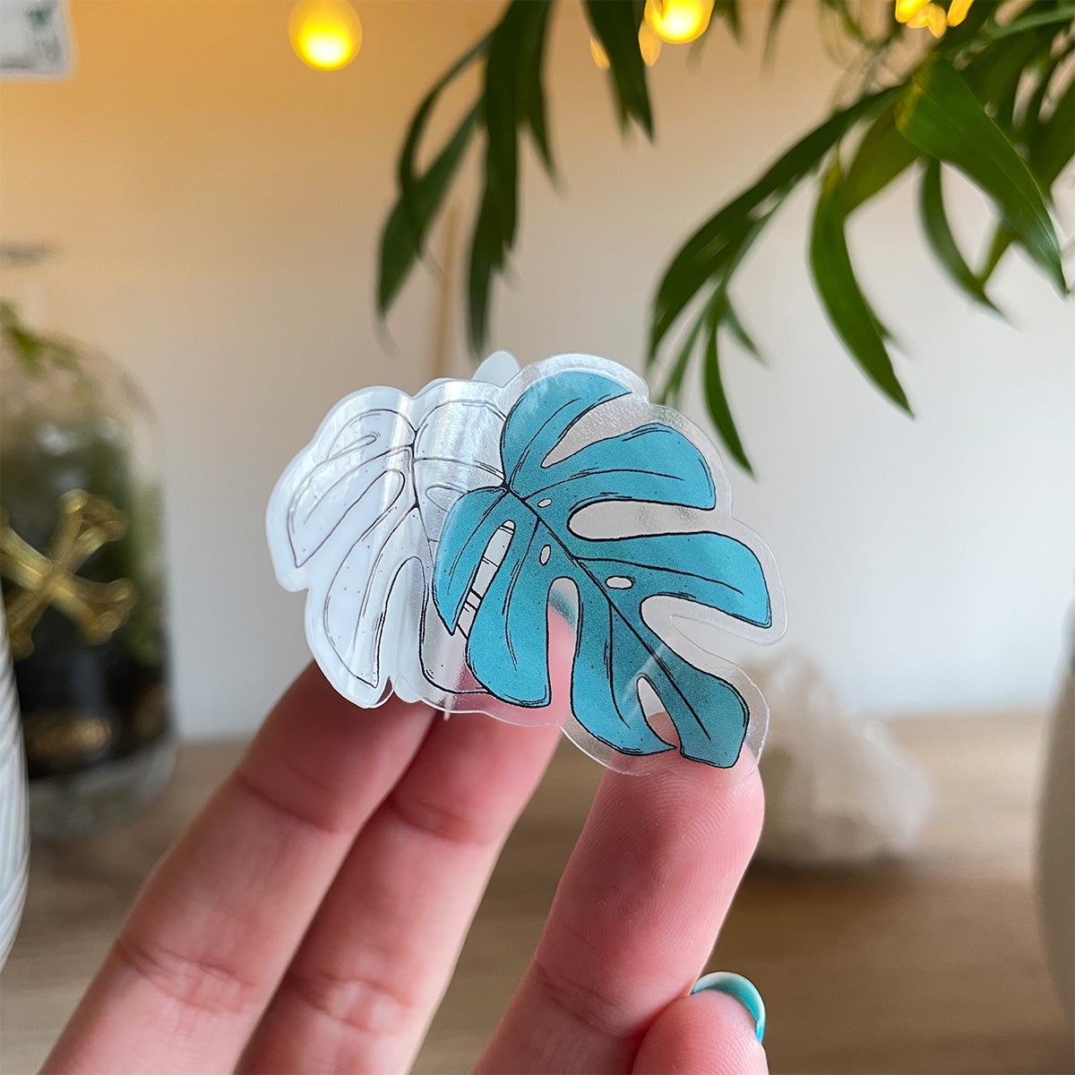 Hand holding clear sticker of two monstera leaves, one is outlined in black, the other teal coloured