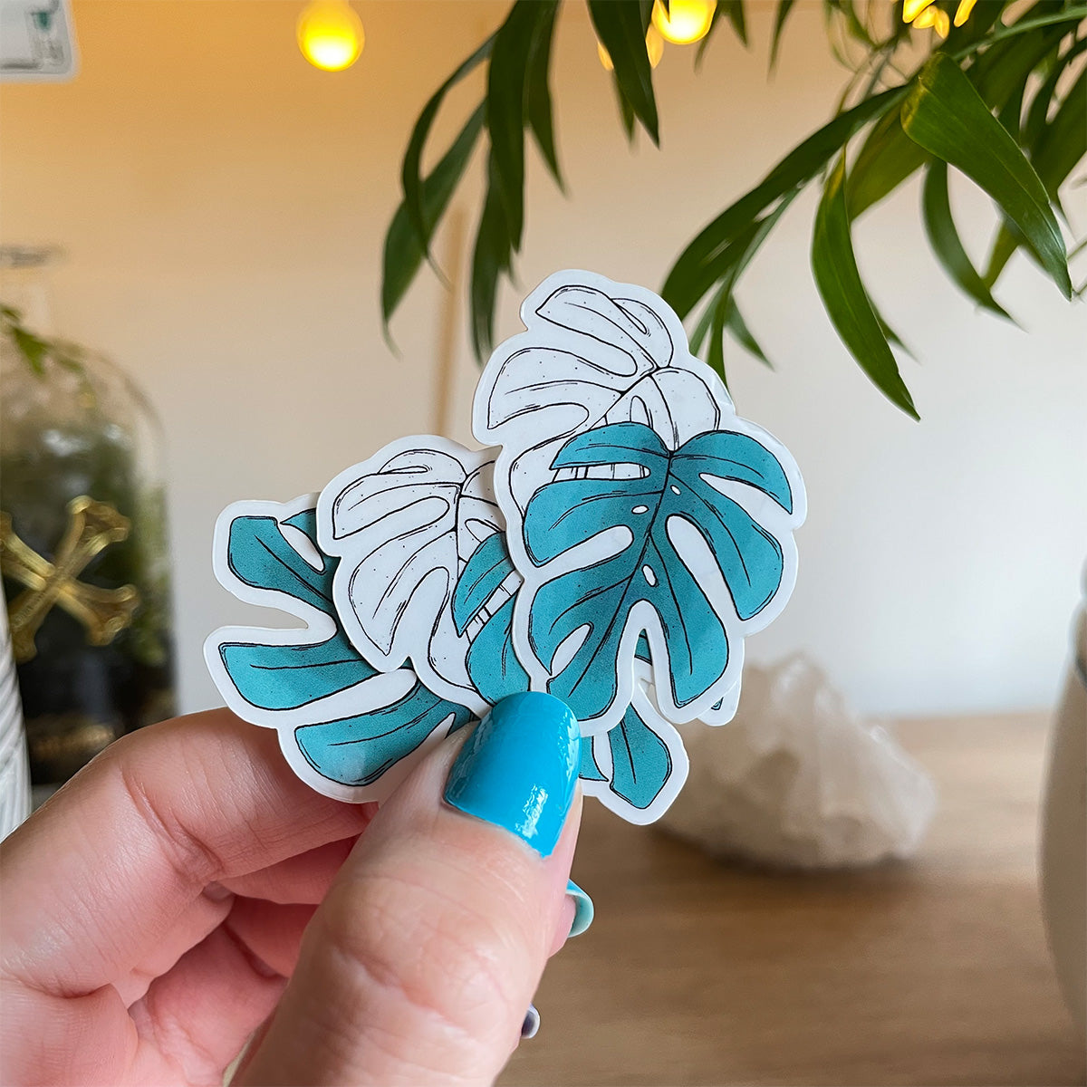 Hand holding several stickers of two monstera leaves, one is outlined in black, the other teal coloured