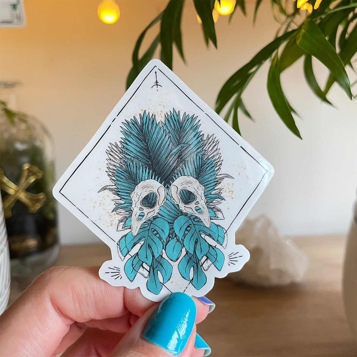 hand holding sticker plant scenario back. Design of tropical teal leaves mirrored with two bird skulls, runes and black lines in diamond shape