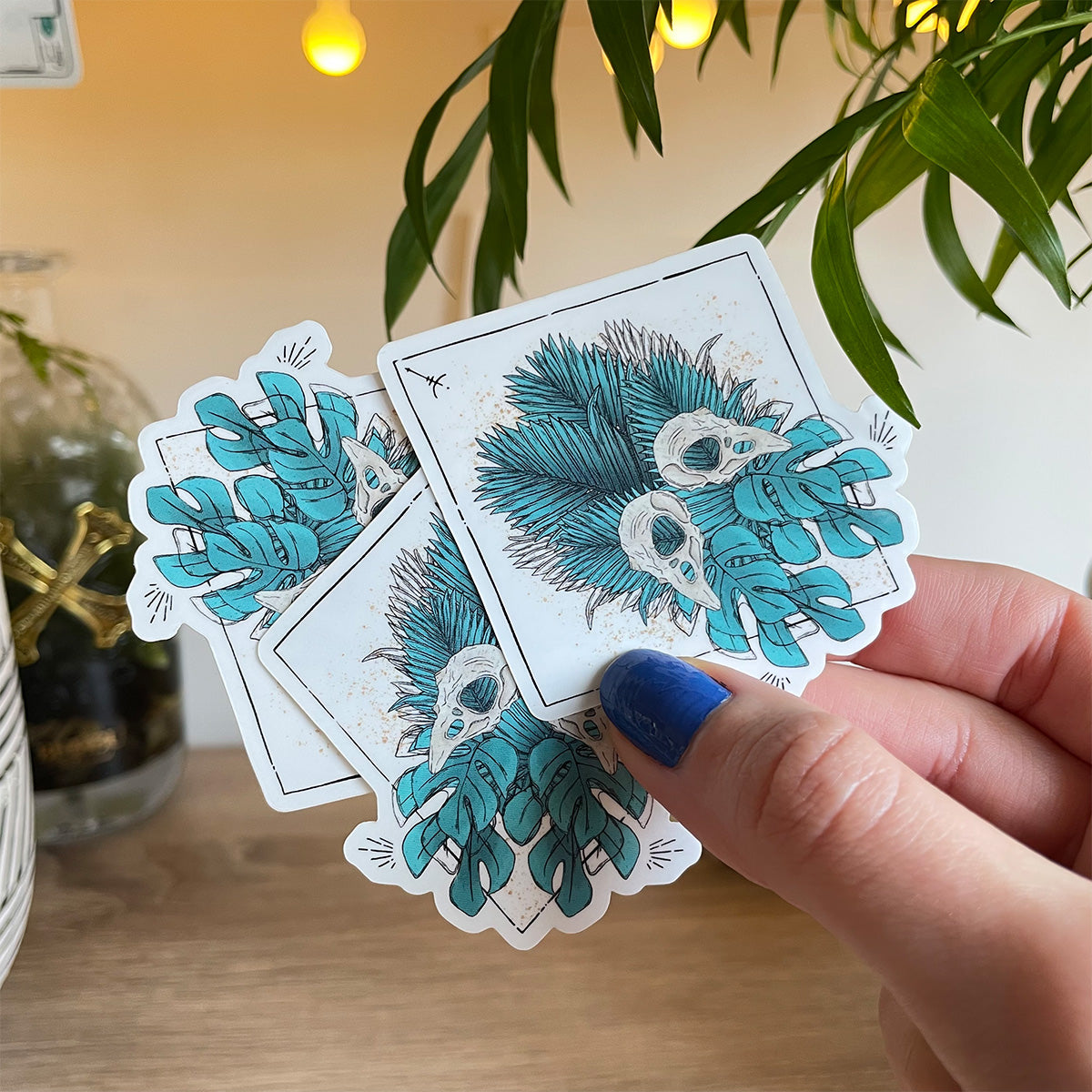 hand holding three stickers plant scenario back. Design of tropical teal leaves mirrored with two bird skulls, runes and black lines in diamond shape