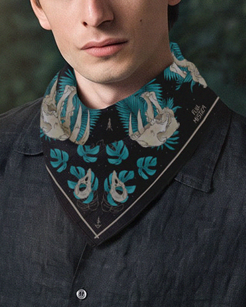 A close-up of a man wearing a printed neck bandana tied diagonally. The neck tie is black with teal tropical leaves and animal skulls of crows and saber tooth tigers