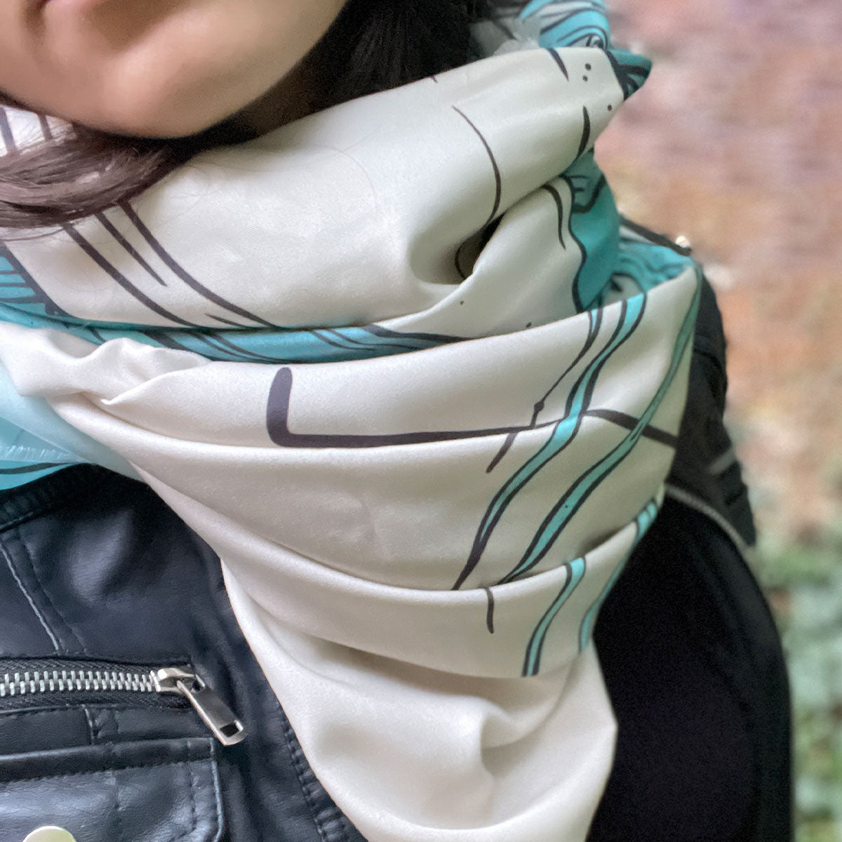 Woman outfit black leather jacket with scarf. Print of plants in teal, cream, black, outdoors.