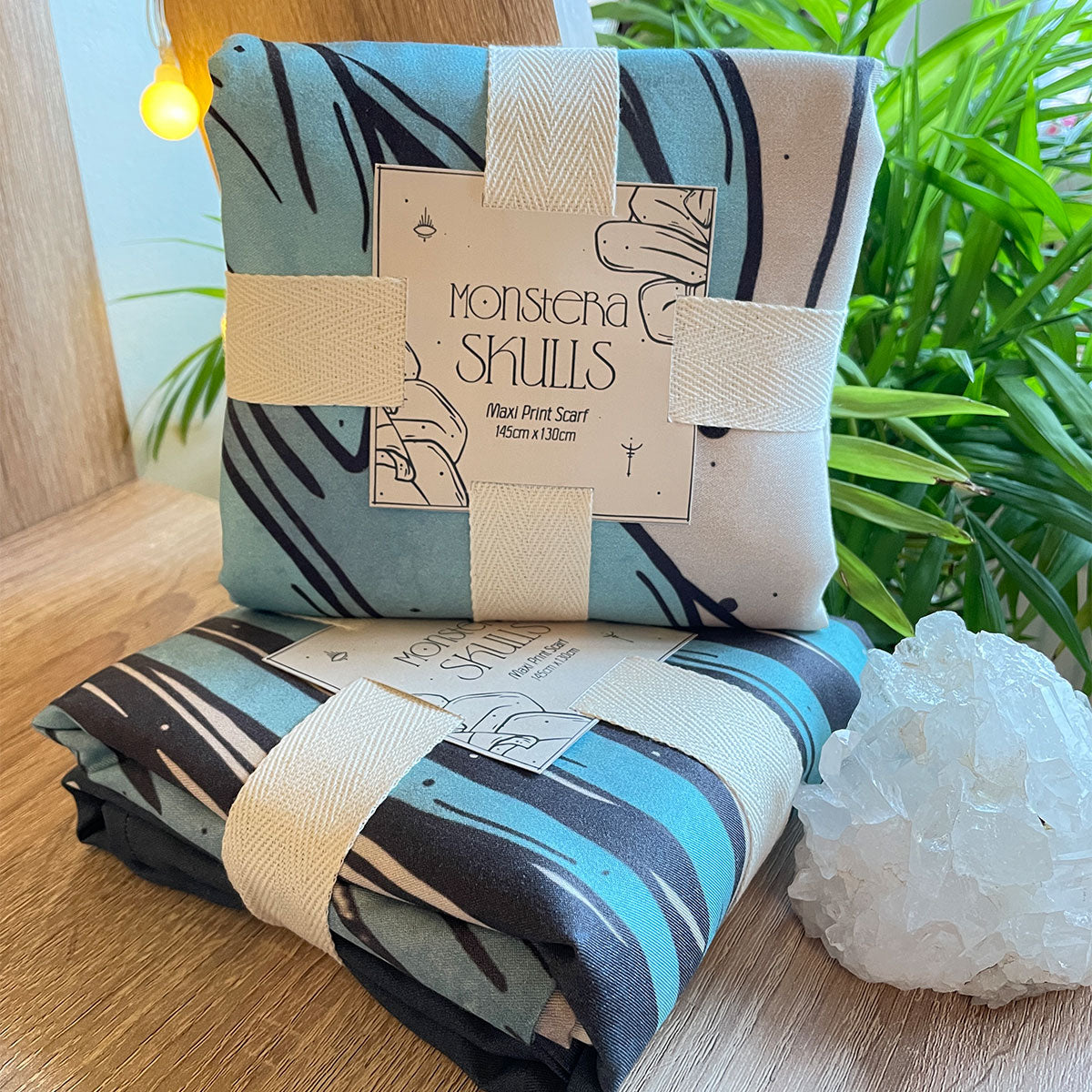 Two folded fabrics in squares, sitting on table, tied with cotton ribbon and paper square with description. monstera skulls tag, teal black and cream colours