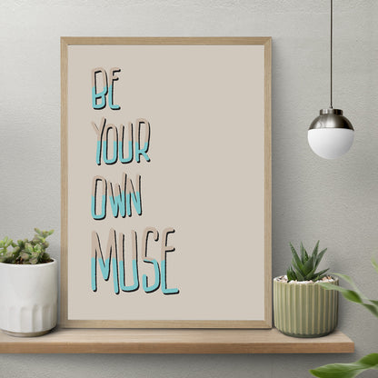 Photo of art print framed sitting on wooden shelf. Print says Be Your Own Muse in tall handwritten letters in cream and turquoise, light cream background 