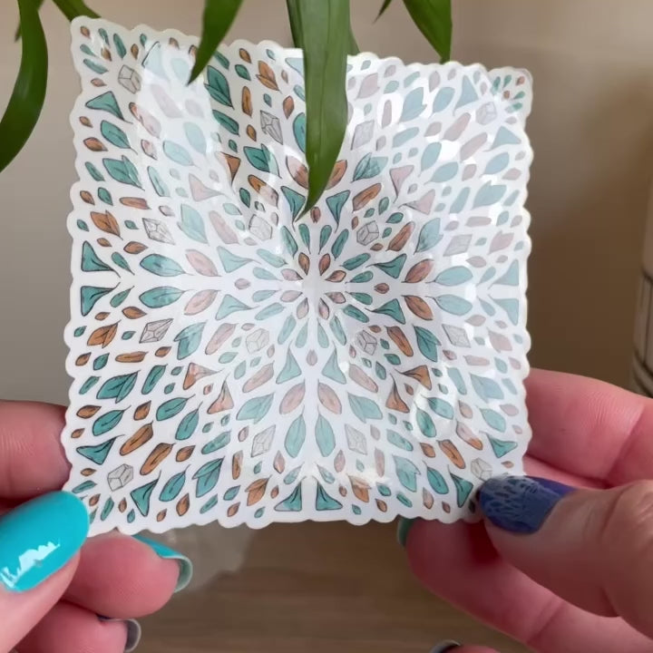 video of hand showing sticker of several tiny leaves and small gemstones in teal and orange organised like a tile shaped like a square