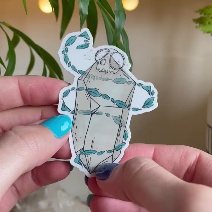 video of hand showing sticker of big quartz crystal with tiny monkey skull on top, swirls of teal leaves floating around it