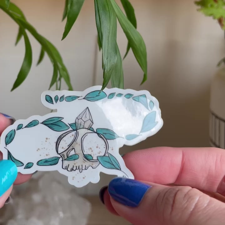 video of hand holding sticker of tiny skull with crystal and several leaves floating around. Background has two plant pots and citrines sitting on table