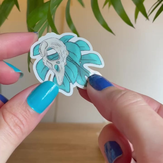 video of hand showing tiny clear sticker of two monstera leaves in teal, bird skull above leaves, big crystal and palm leaves blurry behind