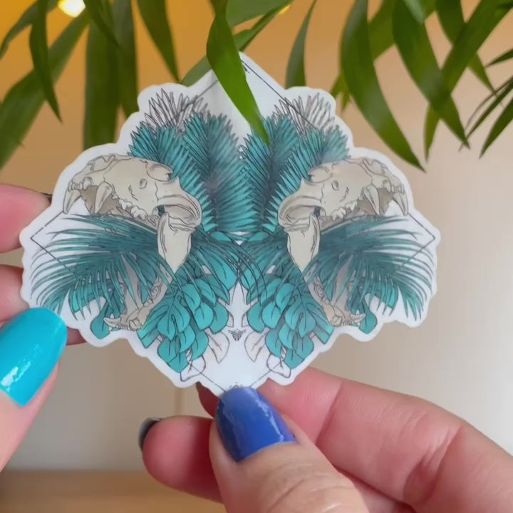 Video of hand holding clear transparent sticker, design of tropical leaves in teal with two lion skulls open mouths mirrored, runes and black lines in wide diamond shape